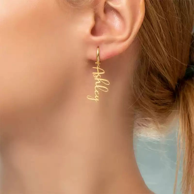 Gold hoop earring with 'Ashley' engraving worn by a person. Stainless Steel Name Jewelry. Custom Gift-Personalized. gift for boyfriend / girlfriend. long-distance relationship gift meaningful keepsake. Memory gift. Christmas couple gift. Birthday Gift. Vertical Charm Ear Cuffs