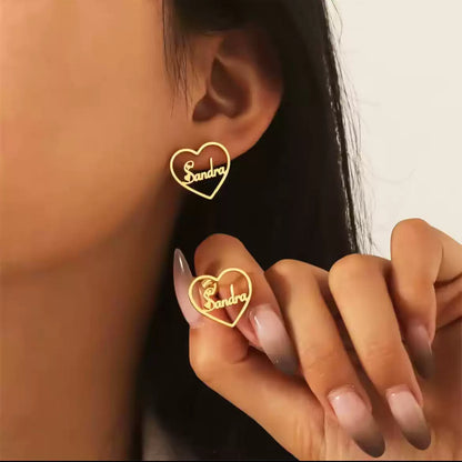 Gold heart-shaped earrings with 'Sandra' engraved, worn by a person.Stainless Steel Name Jewelry. Custom Gift-Personalized. gift for boyfriend / girlfriend. long-distance relationship gift meaningful keepsake. Memory gift. Christmas couple gift. Birthday Gift. Heart Embrace Name Studs 