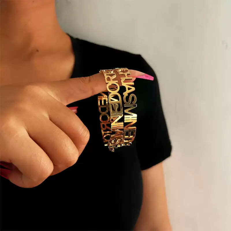 Person wearing a black t-shirt with gold text and pointing to it. Stainless Steel Name Jewelry. Custom Gift-Personalized. gift for boyfriend / girlfriend. long-distance relationship gift meaningful keepsake. Memory gift. Christmas couple gift. Birthday Gift. Statement Loop Name Earrings