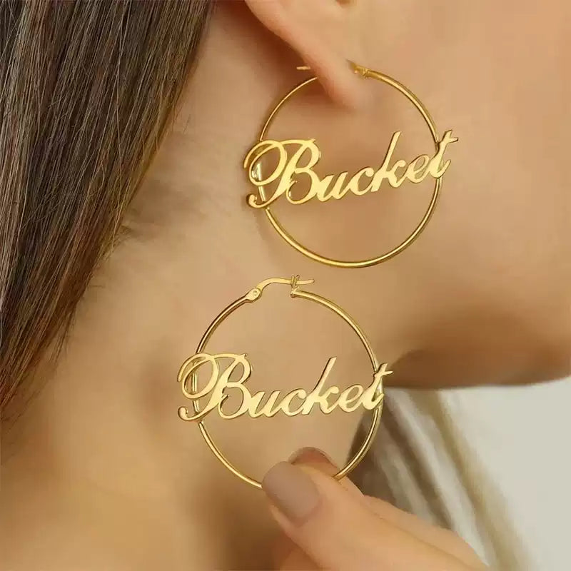 Gold hoop earrings with 'Bucket' text worn by a person Stainless Steel Name Jewelry. Custom Gift-Personalized. gift for boyfriend / girlfriend. long-distance relationship gift meaningful keepsake. Memory gift. Christmas couple gift. Birthday Gift. Retro Classic Hoop Name Earrings
