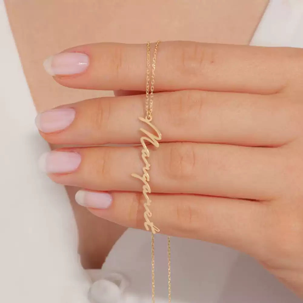 Gold 'Marquesas' necklace on a hand with pink nail polish against a neutral background Stainless Steel Name Jewelry. Custom Gift-Personalized. gift for boyfriend / girlfriend. long-distance relationship gift meaningful keepsake. Memory gift. Christmas couple gift. Birthday Gift. Vertical Name Necklace
