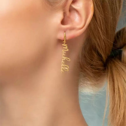 Gold hoop earrings with 'Michelle' engraving worn by a person. Stainless Steel Name Jewelry. Custom Gift-Personalized. gift for boyfriend / girlfriend. long-distance relationship gift meaningful keepsake. Memory gift. Christmas couple gift. Birthday Gift. Vertical Charm Ear Cuffs