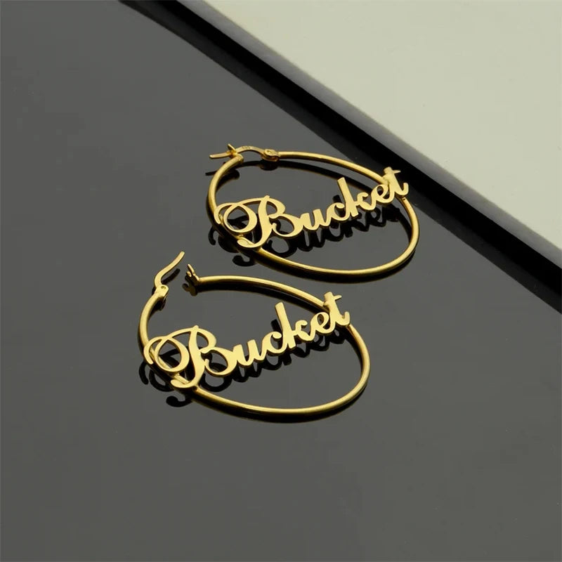 Gold hoop earrings with 'Bucket' text on a reflective surface Stainless Steel Name Jewelry. Custom Gift-Personalized. gift for boyfriend / girlfriend. long-distance relationship gift meaningful keepsake. Memory gift. Christmas couple gift. Birthday Gift. Retro Classic Hoop Name Earrings