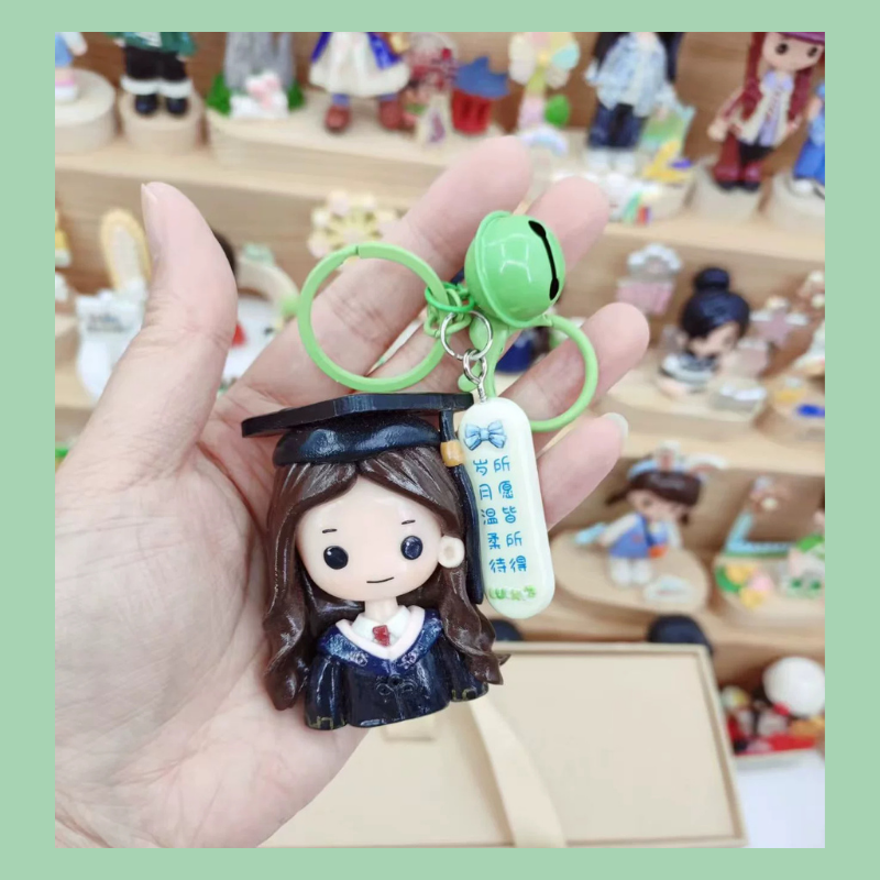 Graduation-themed keychain with a character wearing a cap and gown, held in a hand with a blurred background of figurines. Personalized couple present. Gift for boyfriend girlfriend. Custom-made. Personalized. Perfect gift idea. Meaningful keepsake. Wedding gift. Anniversary gift. Birthday gift